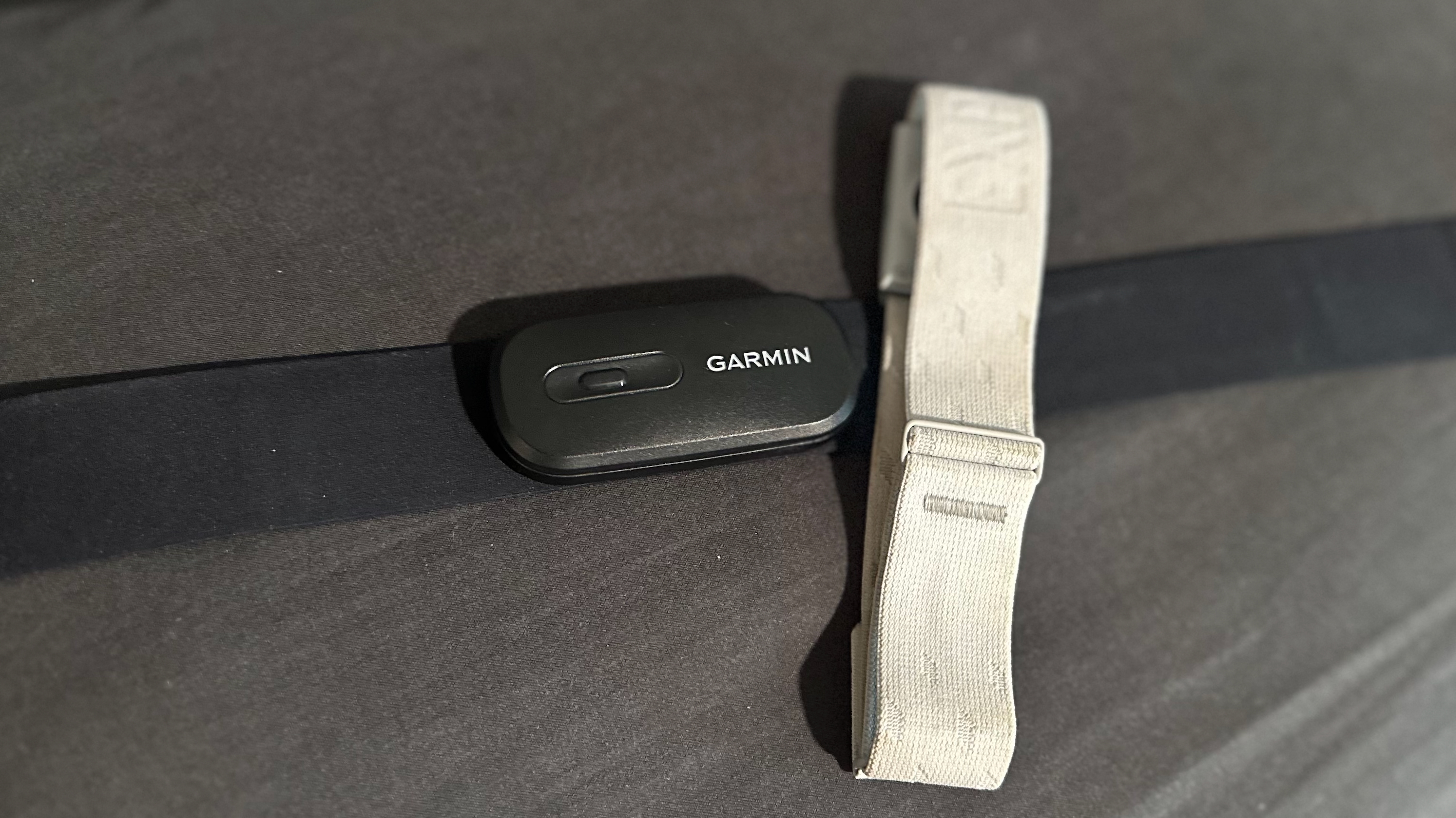 A Garmin HRM 200 chest strap laying on a pillow, with the COROS HRM armband laying perpendicular atop its right side.